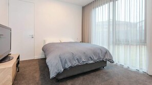 Ett hotellrum med en säng, en tv på ett stativ och ett stort fönster med gardiner.