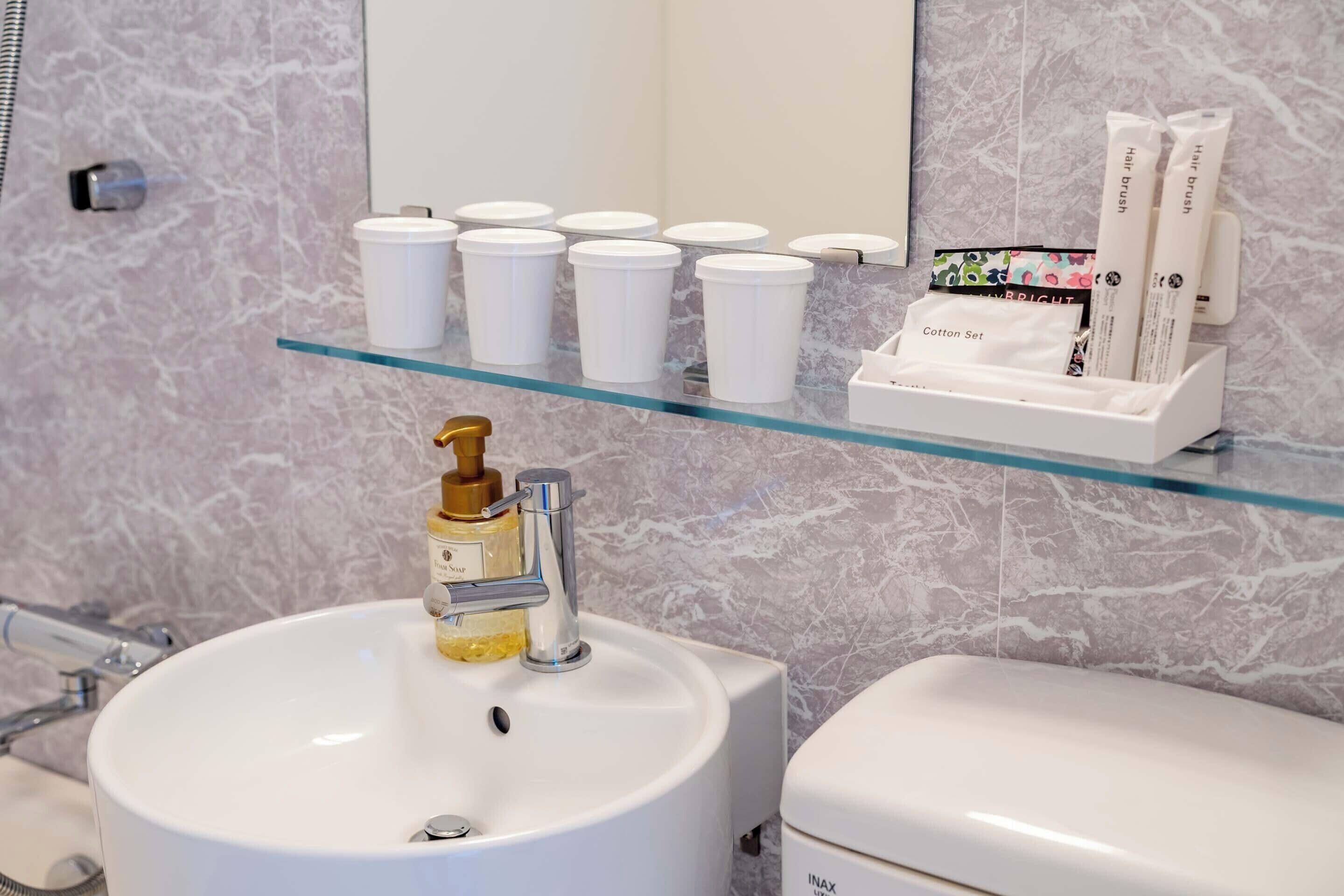 Casa de banho com parede de mármore, um prateleira de vidro com produtos de higiene pessoal, uma pia branca com dosador dourado e uma sanita branca.
