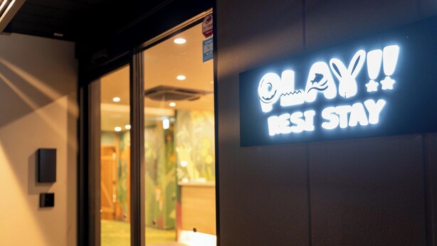 A sign with the text "PLAY!! RESI STAY" in a well-lit indoor setting.