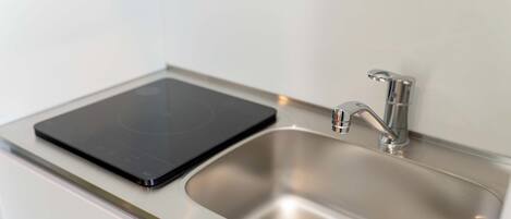 Un évier en acier inoxydable, doté d’un robinet moderne et d’une plaque de cuisson à induction noire, le tout installé sur un plan de travail blanc.