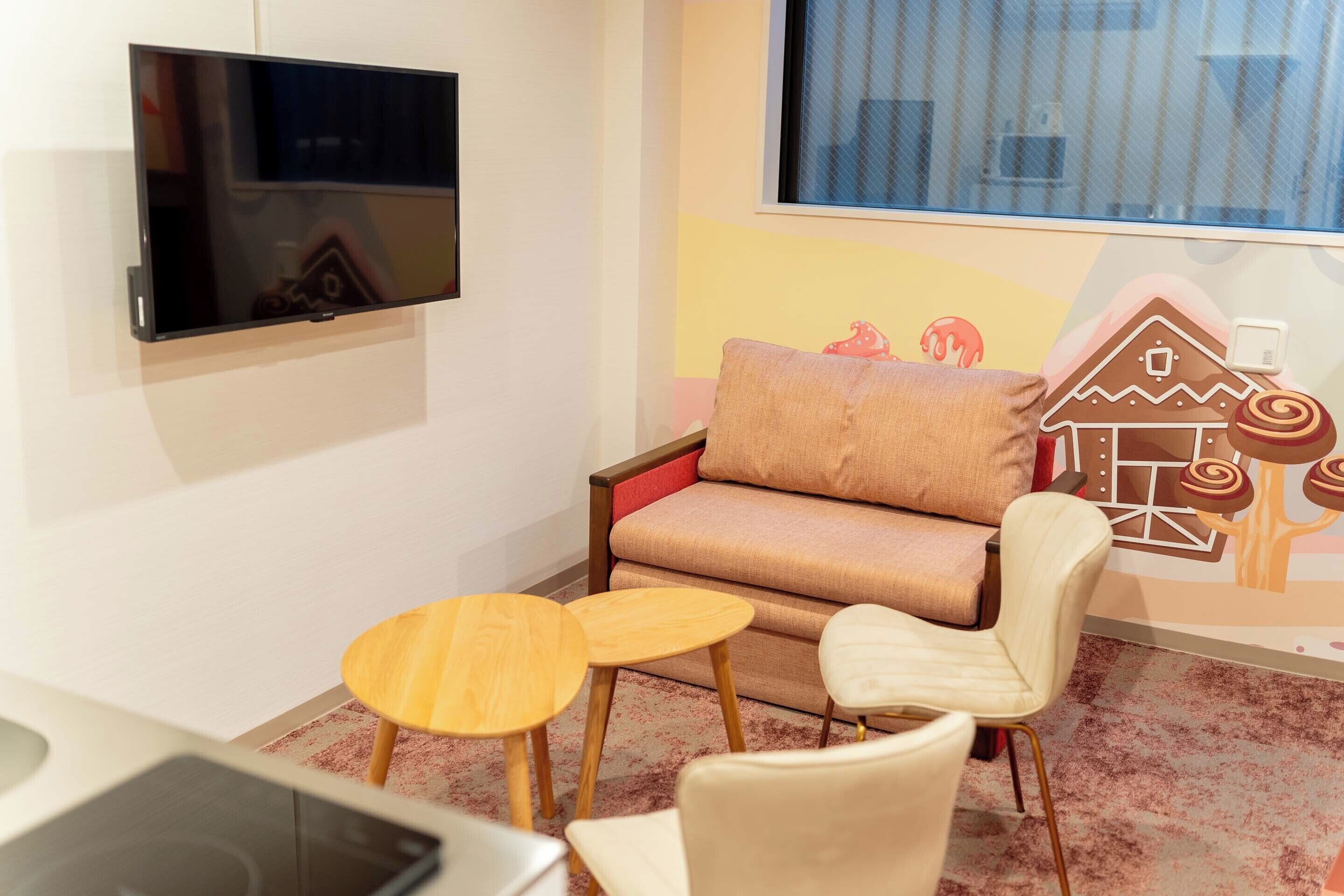 A cozy room with a flat-screen TV mounted on the wall, a sofa with a cushion, two small wooden tables, and a chair.