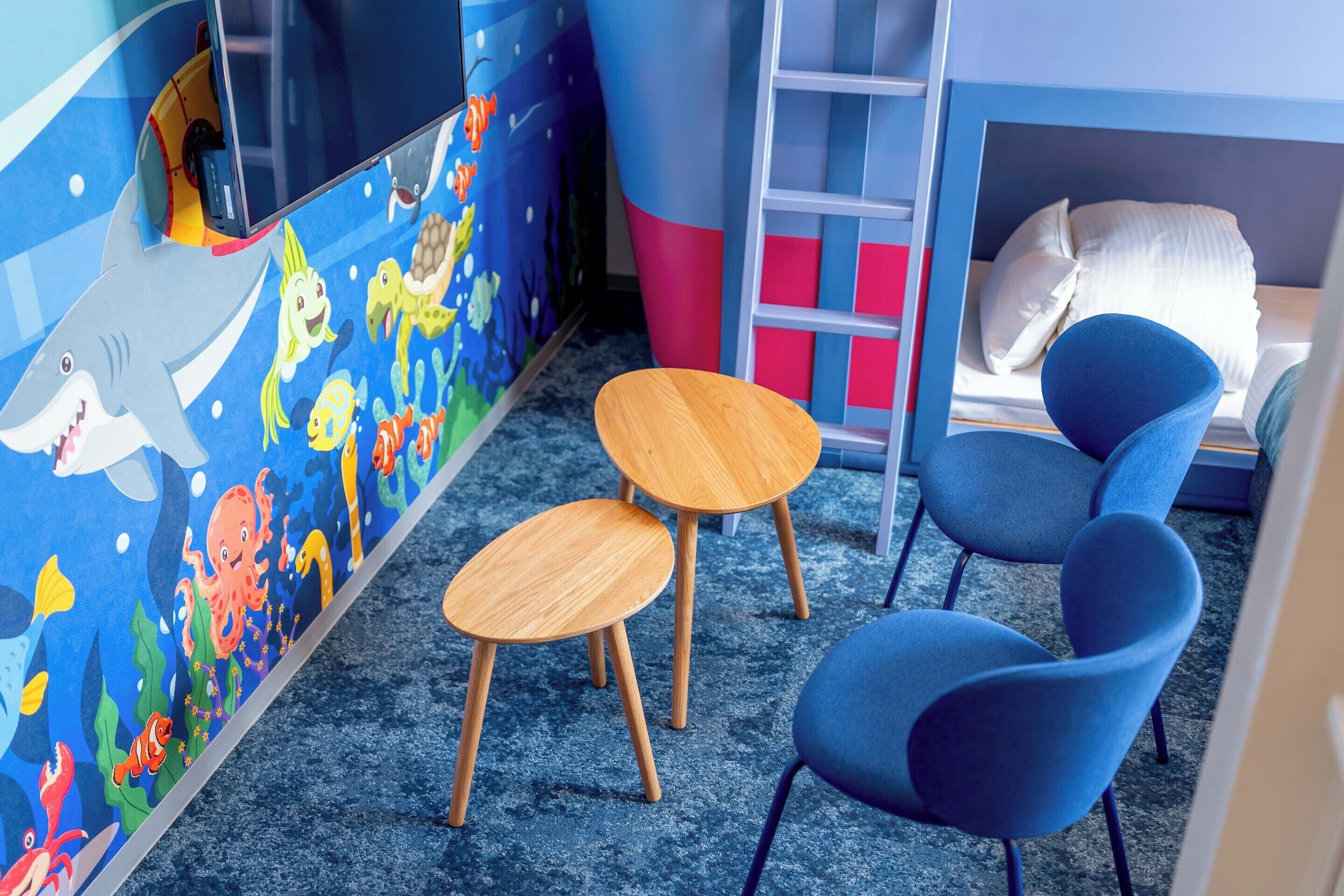 Un salón infantil con un mural temático de océano azul, dos mesas de madera y sillas azules.
