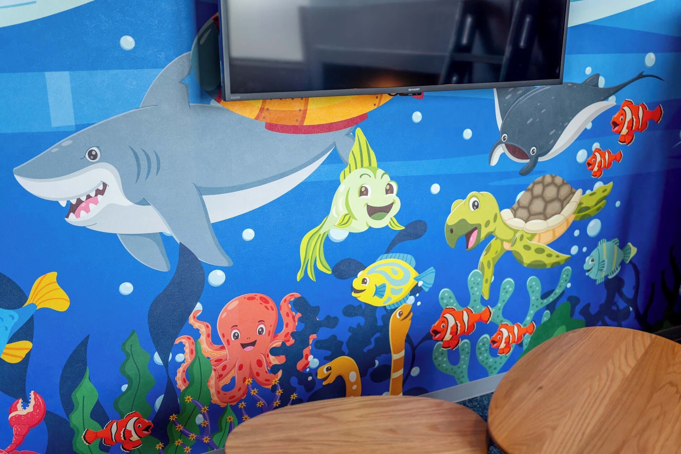 A room with a colorful underwater mural, a television mounted on the wall, and a wooden chair.