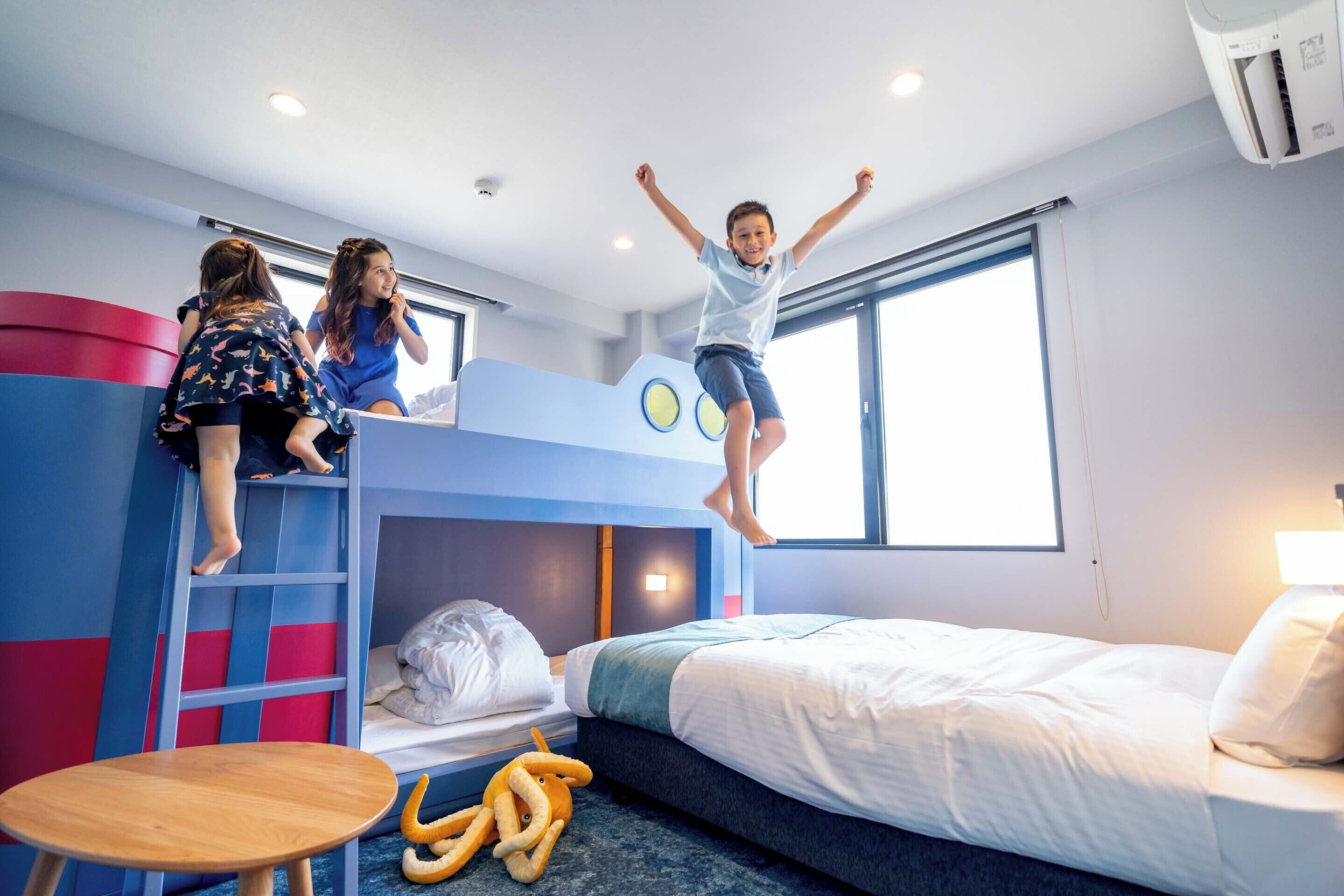 A child jumping off a bunk bed in a room with a bed, a table, and a window.