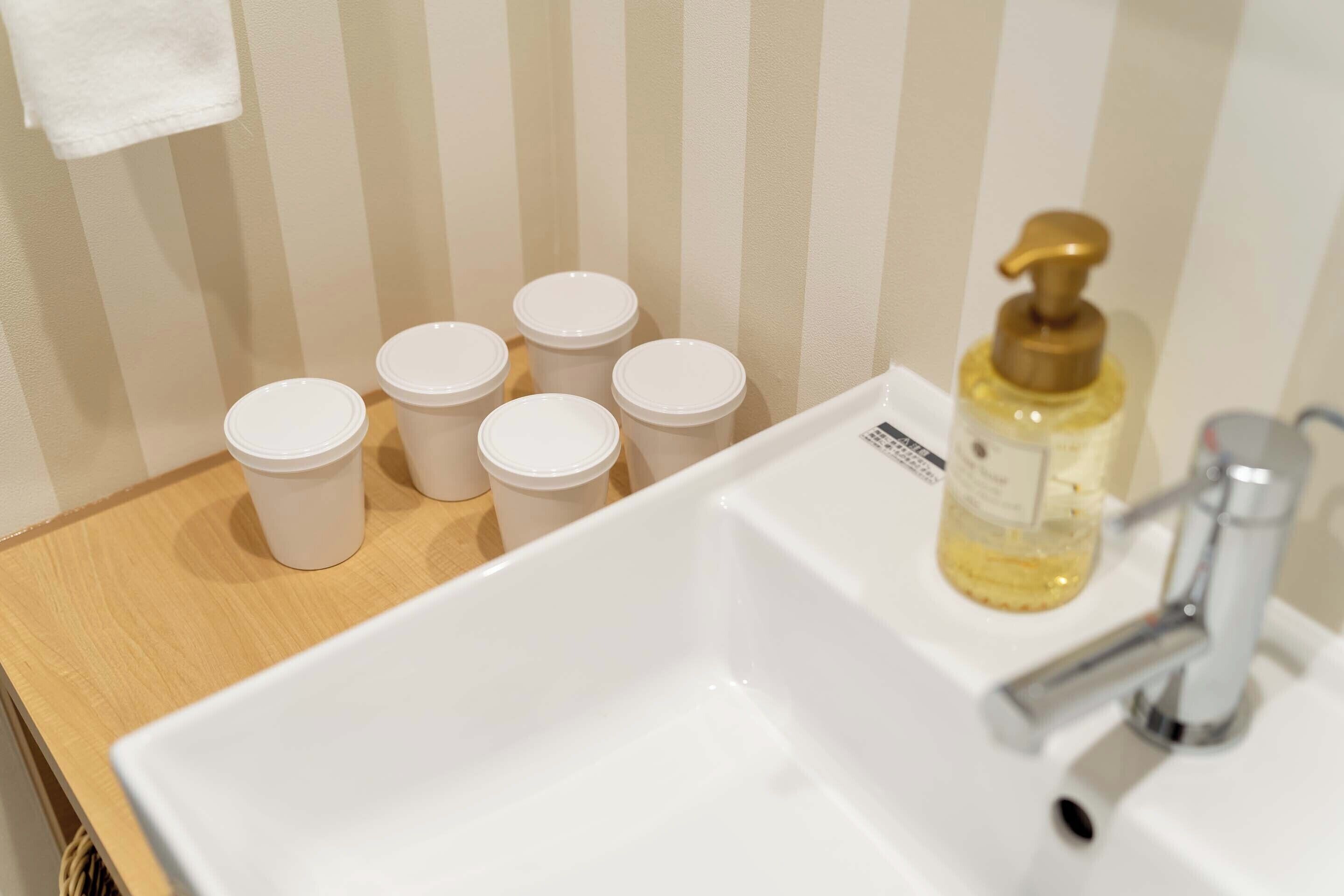 A bathroom with a white sink, a bottle of soap, and five white cups on a wooden shelf.