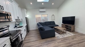 A modern kitchen with white cabinets, a stainless steel oven, and a coffee maker. There is a gray sectional sofa, a wooden coffee table, and a TV on a wooden stand.