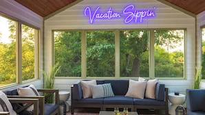 A cozy seating area with a dark blue sofa, two armchairs, and a coffee table with fruits and glasses. A neon sign reads "Vacation Sippin'". - Galena Getaway: Sauna, Hot Tub, Fire Pit, and Arcade Fun Awaits! (Galena)