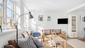 Een woonkamer met een bruine leren bank, gestreepte kussens, een houten salontafel en een vloerlamp. Er is een televisie aan de muur bevestigd en een kast met glazen deuren.