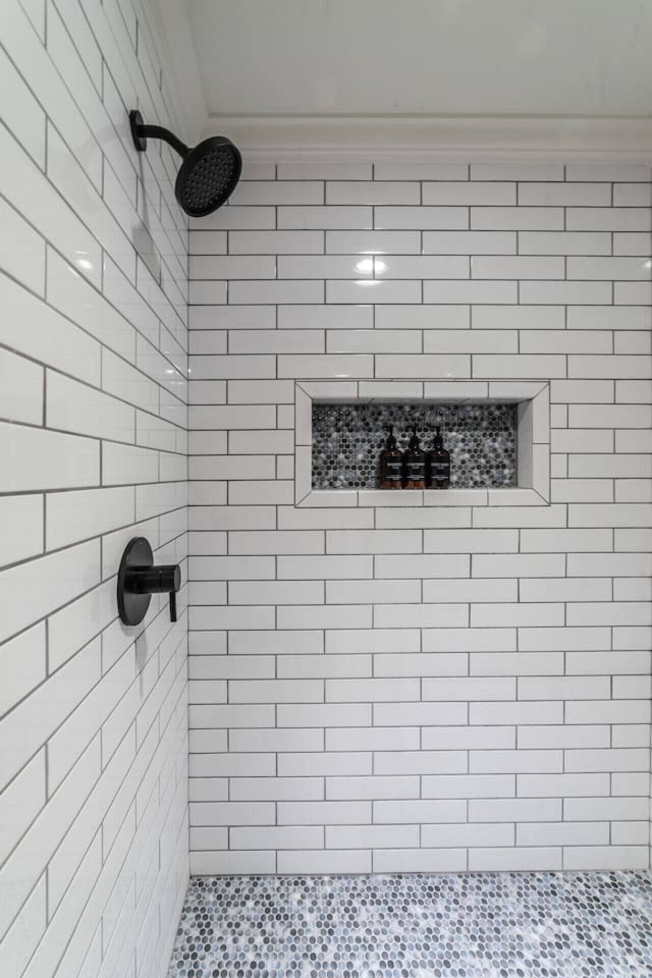 Et brusebad med hvide metrofliser, et sort brusehoved og en lille hylde med flasker.