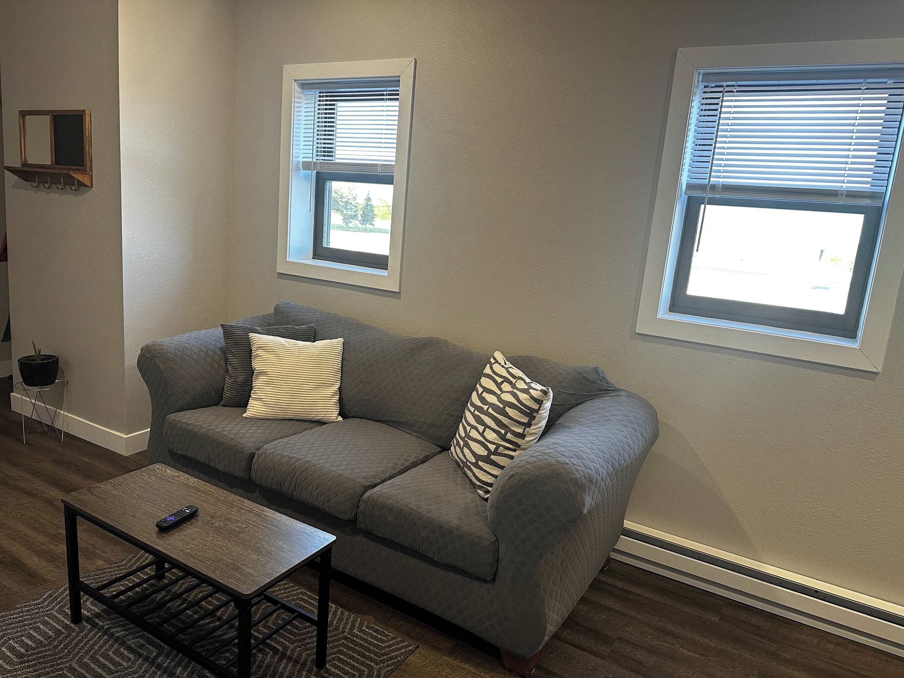 A living room with a gray sofa, two windows with blinds, a small table, and a potted plant.