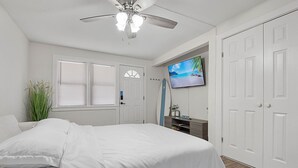 Ein Schlafzimmer mit Bett, Deckenventilator, Fernseher, einer Tür und einem Fenster mit Jalousien.
