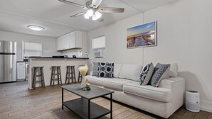 Eine moderne Küche mit Kühlschrank, Herd und Barhocker. Es gibt einen Deckenventilator, einen Esstisch und eine Couch mit gemusterten Kissen.
