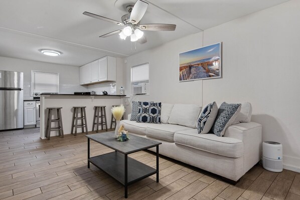 A modern kitchen with a refrigerator, stove, and bar stools. There is a ceiling fan, a dining table, and a sofa with patterned cushions.