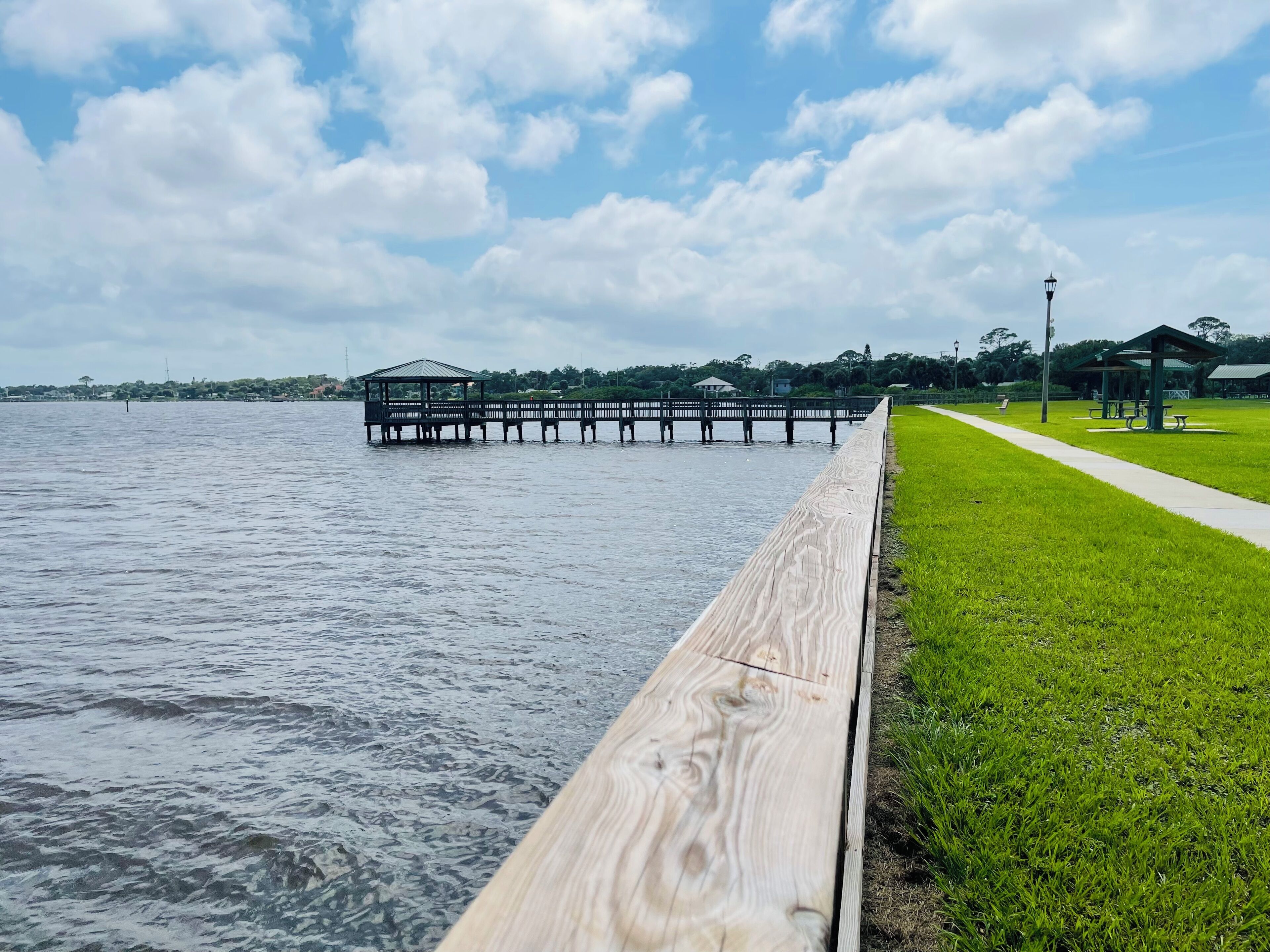 Un paseo marítimo de madera bordeando un lago, con un pabellón y mesas de picnic.
