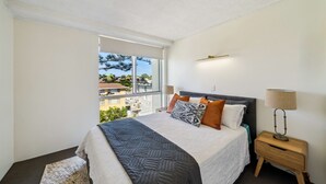 Un dormitorio con una cama grande, una mesita de noche, una lámpara y vistas a una zona residencial por la ventana.