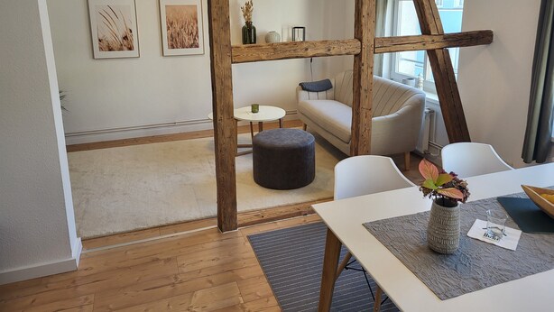 Un interior moderno con estructura de techo de madera, un sofá, una mesa de comedor con un florero y vista a un edificio exterior.