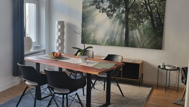 Un comedor con una mesa de madera, sillas negras, una planta en maceta y un cuadro grande con motivos forestales en la pared.