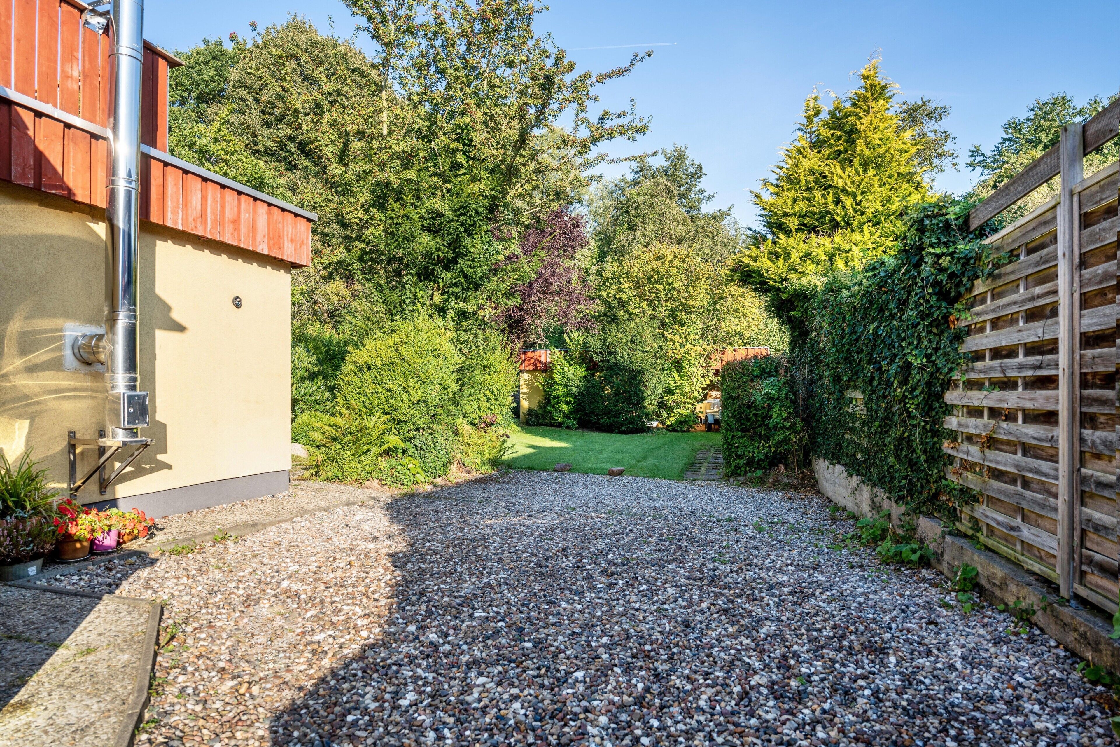 Ein Schotterweg führt zu einem Haus mit Holzzaun und einem Garten mit verschiedenen Pflanzen.