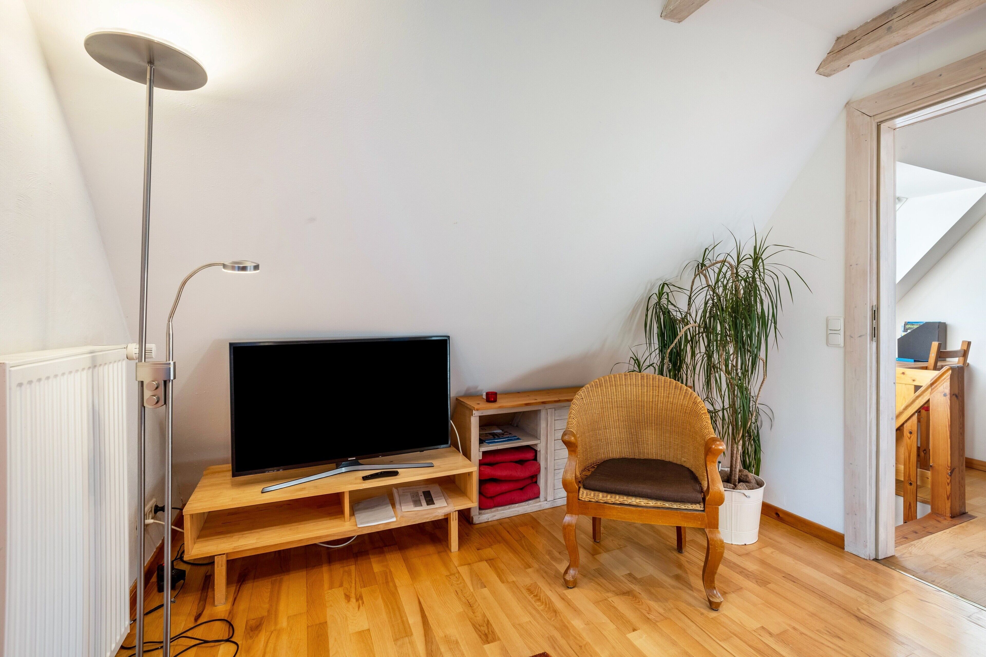 Ein Wohnzimmer mit Fernseher auf Holzfuß, einer Stehlampe, einem Sessel, einem Blumentopf und einer Treppe, die in ein weiteres Zimmer führt.