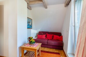 Une petite pièce avec un canapé rouge, une table en bois recouverte d’une nappe à carreaux et un tableau accroché au mur.