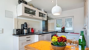 Cocina compacta con armarios blancos, cafetera, hervidor y mesa de comedor con un mantel individual amarillo.