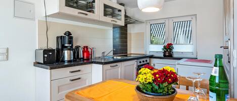 Cocina compacta con armarios blancos, cafetera, hervidor y mesa de comedor con un mantel individual amarillo.