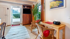 Una habitación con puerta de madera, una silla de mimbre, una mesa con cojín rojo, una planta y un cuadro en la pared.