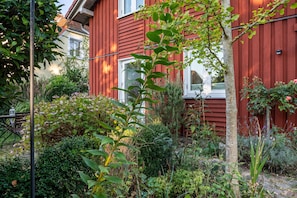 A red wooden house with a garden in front, a bench, and a tree.