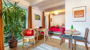 Una habitación con un ventanal grande, una planta en maceta, mobiliario de mimbre, una cama con ropa de cama roja y una mesa con una botella y vasos.