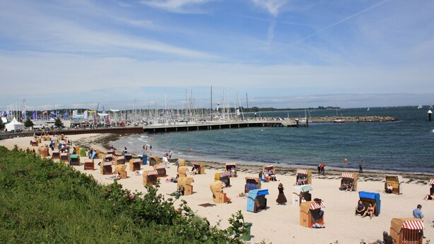 A beach with numerous beach huts and a pier with sailboats.