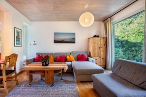 Ein modernes Wohnzimmer mit Holzboden, einem großen Fenster mit Blick auf Grünflächen, einem grauen Sofa, einem Holzschrank und einem Tisch mit bunten Kissen.
