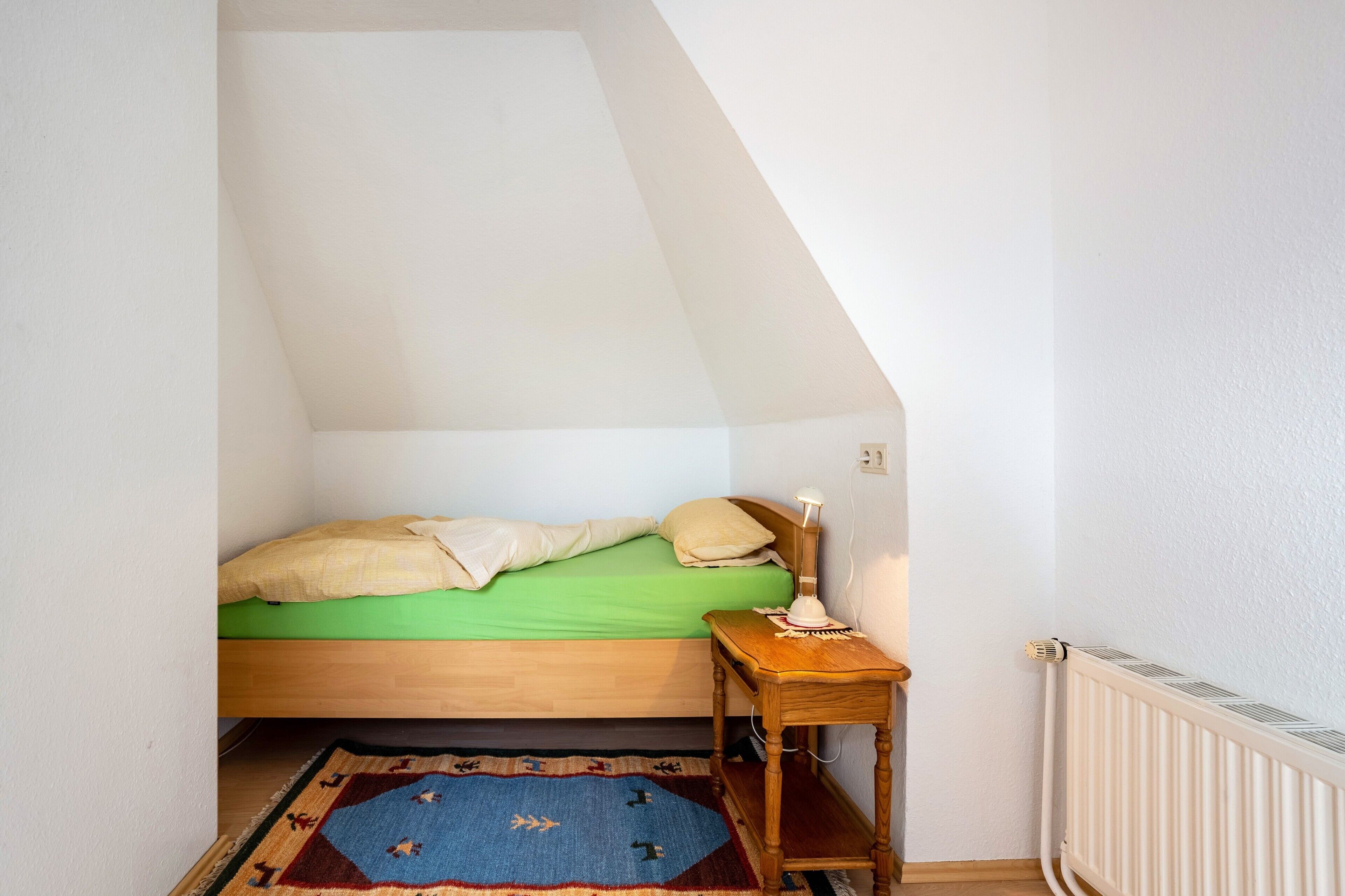 A small bedroom with a single bed, a nightstand, a radiator, and a patterned rug.