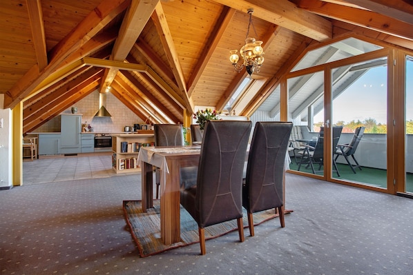 A spacious dining area with a high ceiling and wooden beams, a kitchen in the background, and large sliding glass doors to an outdoor patio.