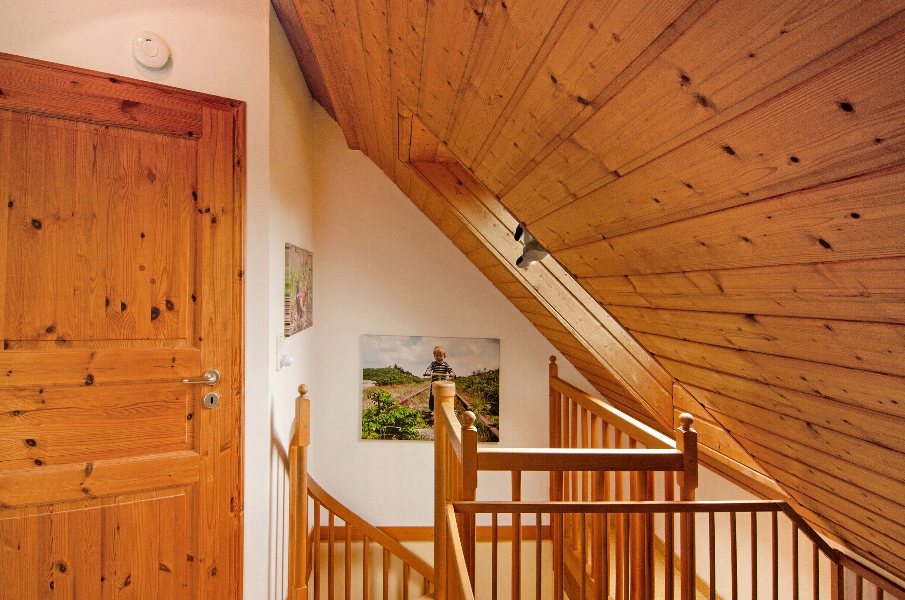 Eine Holztreppe führt zu einem Dachboden mit geneigter Decke, einer Holztür und einem gerahmten Bild an der Wand.