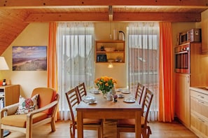 A dining area with a wooden table set for four, surrounded by chairs, a vase of flowers, and a kitchen area with appliances.