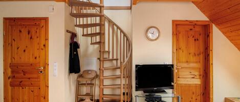Una escalera de madera que lleva a un altillo, un televisor sobre un pedestal de vidrio y puertas de madera.