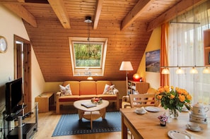 A cozy room with a wooden ceiling, a sofa, a coffee table, a TV, and a window with a view of greenery.