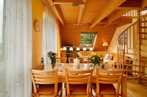 A wooden dining table with chairs, a clock on the wall, and a window with a view of trees.