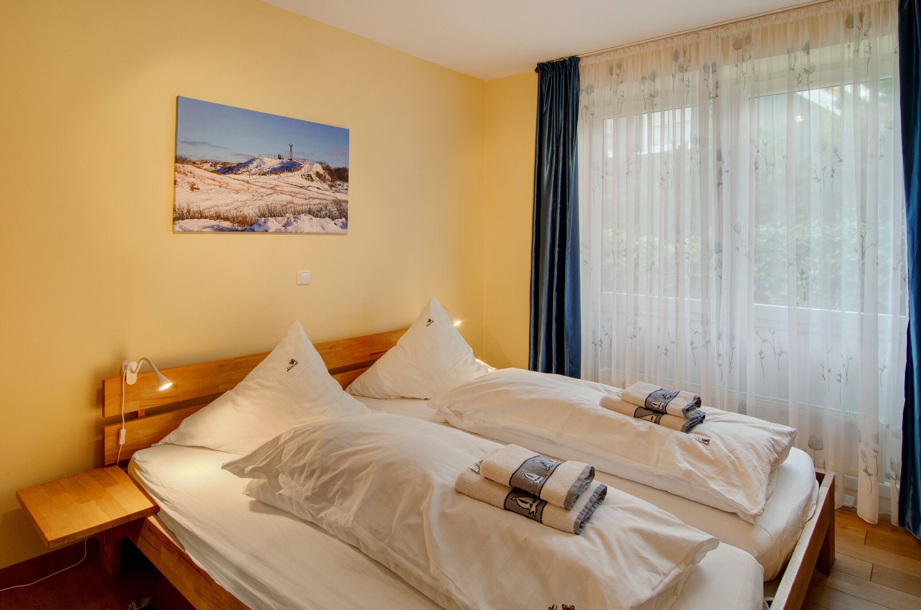Ein Doppelbett mit weißen Leinen, ein Nachttisch mit Lampe, ein Gemälde an der Wand und durchsichtige Vorhänge.