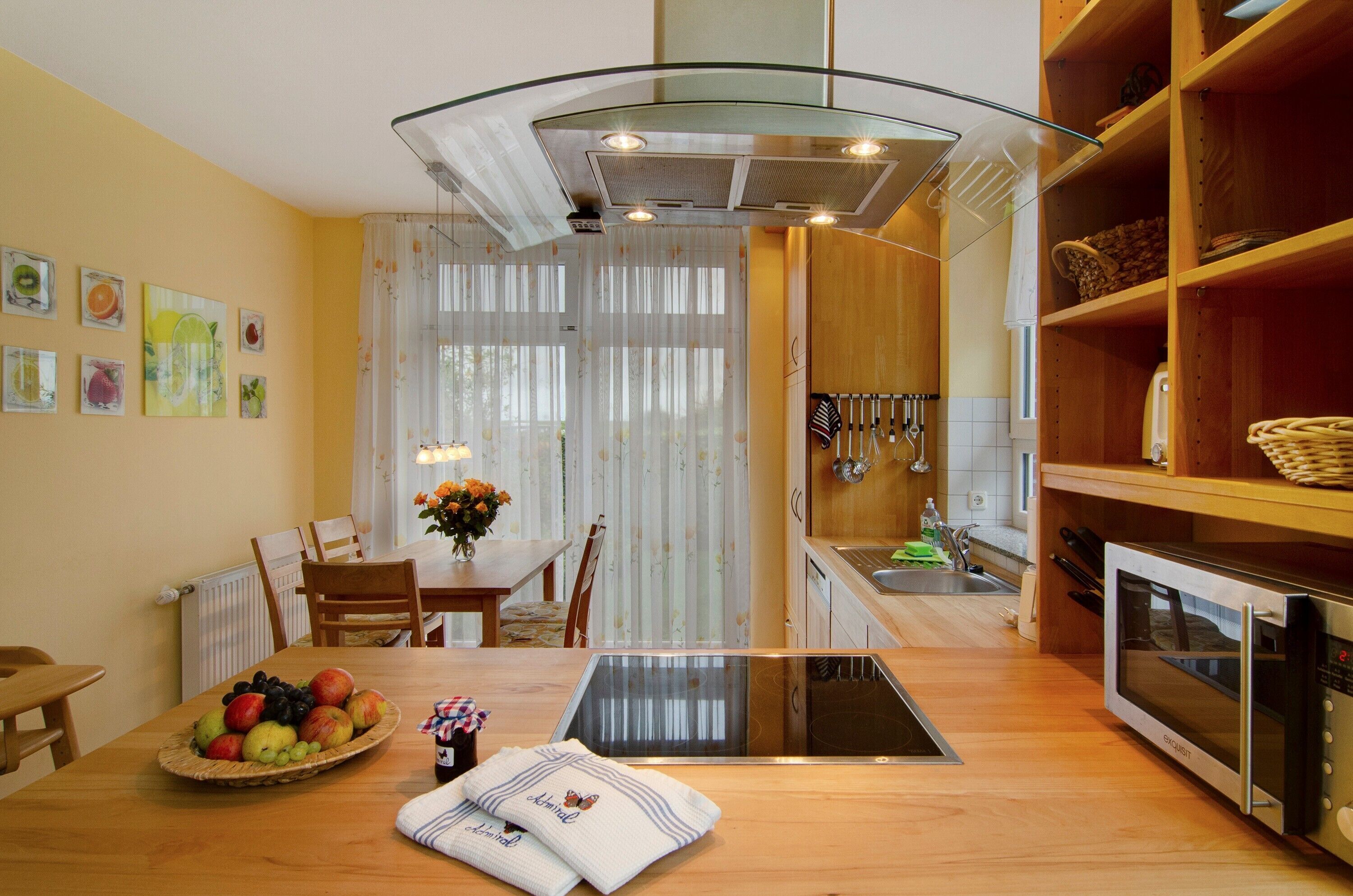 Een keuken met een eettafel, een fruitschaal, een magnetron en een raam met gordijnen.