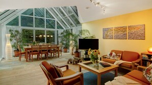 Amplia sala de estar con techo de vidrio, muebles de madera, televisor de pantalla plana y varias plantas en macetas.
