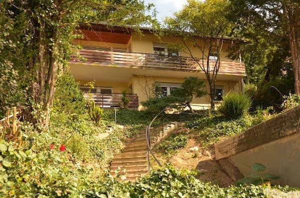 Un bâtiment à deux étages doté d’un balcon, entouré de verdure et agrémenté d’un escalier en bois.