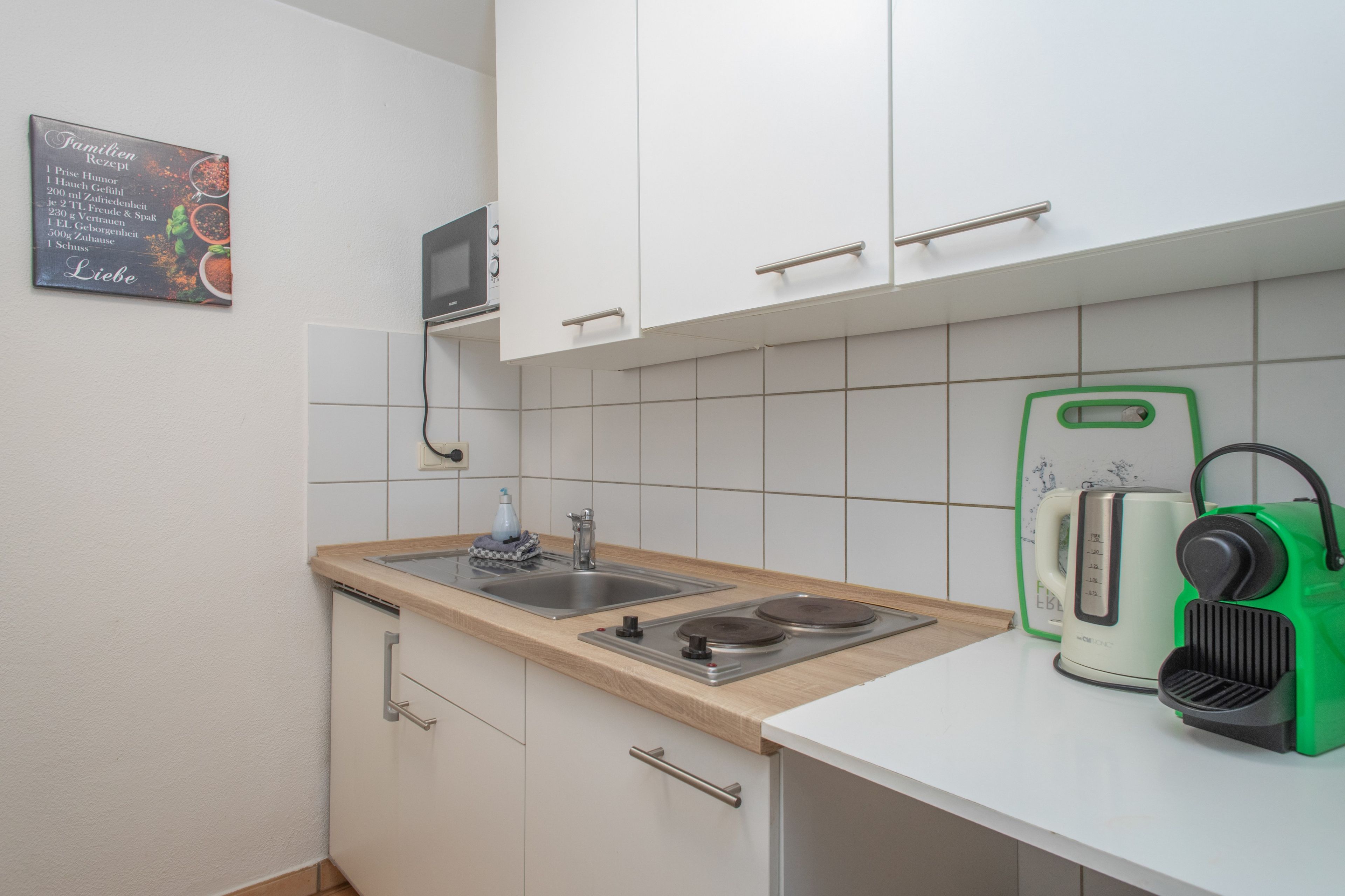 A compact kitchen with white cabinets, a stainless steel sink, a microwave, and a kettle.