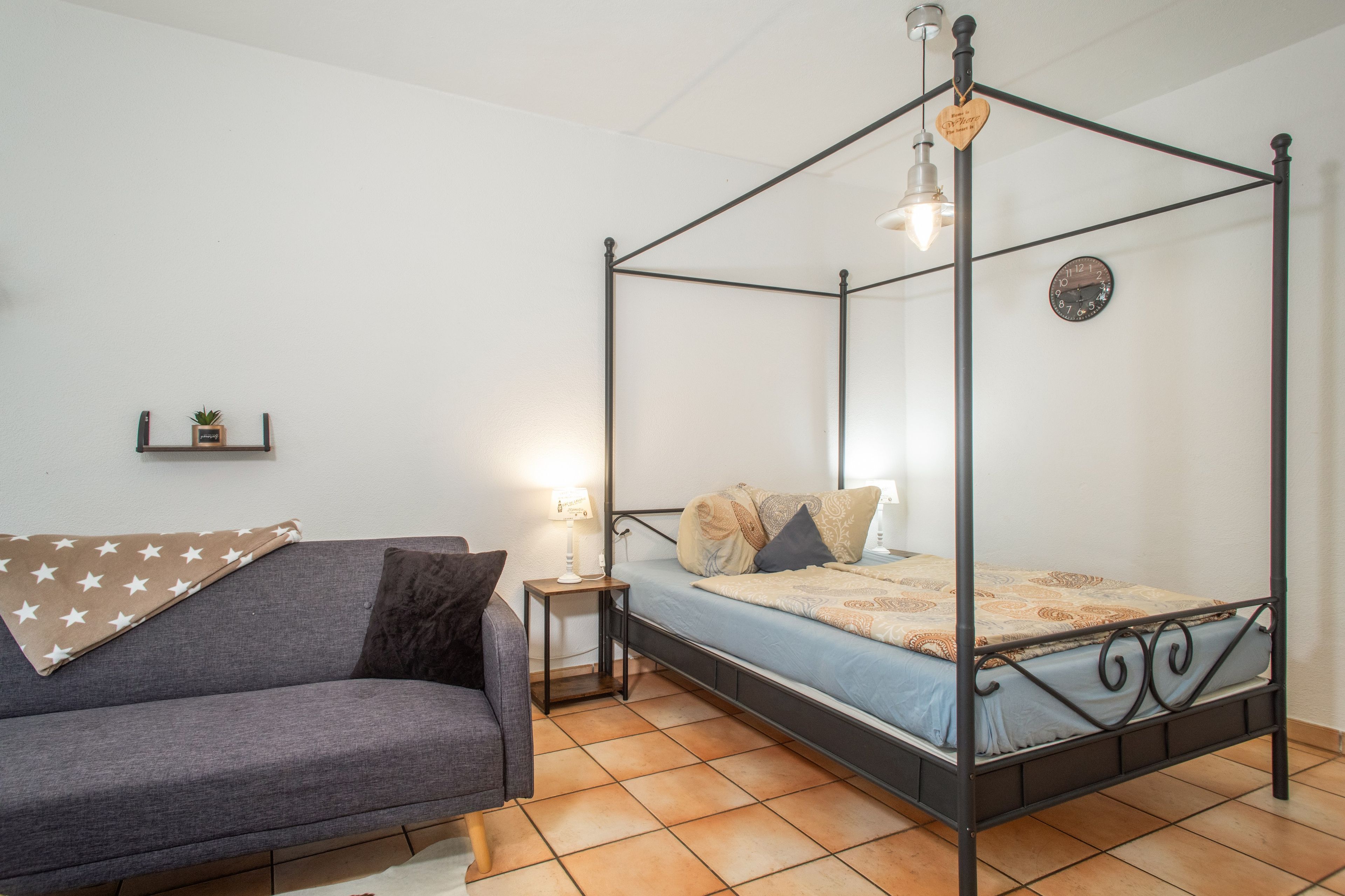 Un letto a baldacchino con una lampada sopra, un comodino con abat-jour, un divano con una coperta a motivi stellari e una sveglia a muro.