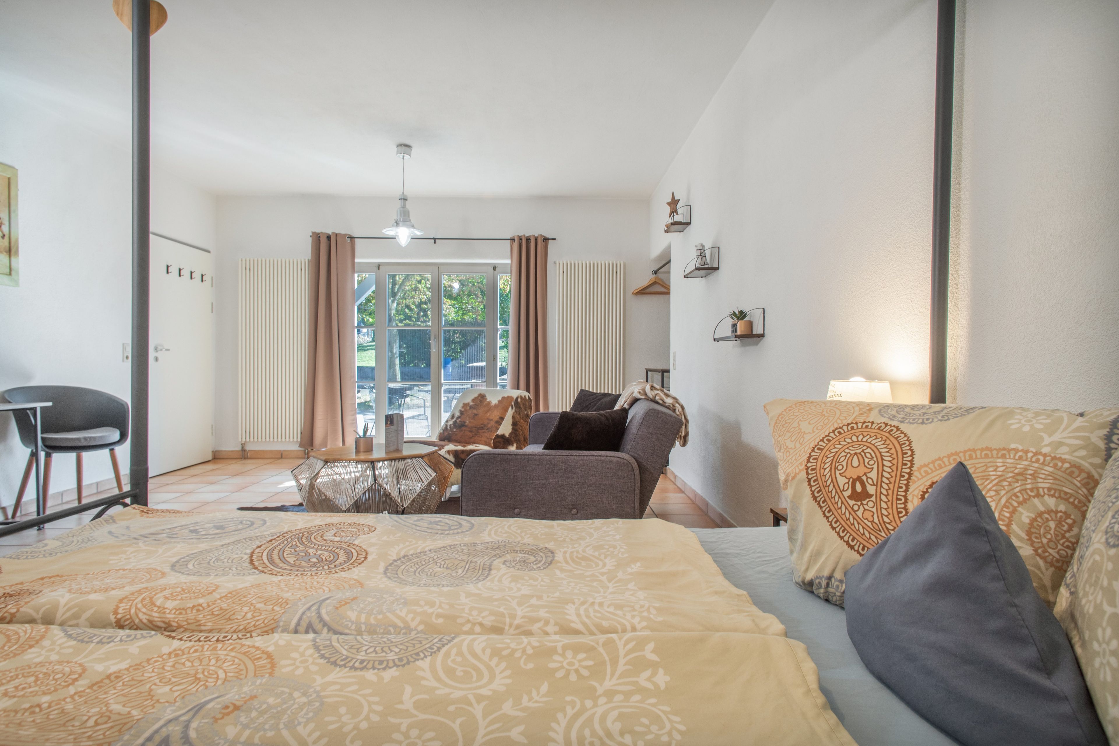 Een hotelkamer met een bed, een bank, een tafel en een stoel. Er is een raam met gordijnen, een radiator en een hanglamp.