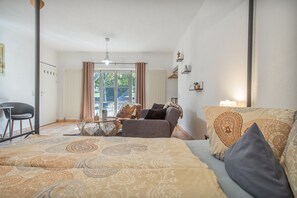 A hotel room with a bed, a sofa, a table, and a chair. There is a window with curtains, a radiator, and a hanging light fixture.