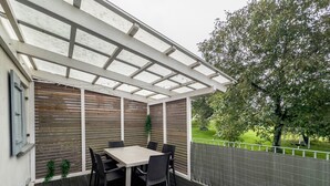 Un patio exterior con mesa y sillas, una pérgola y vistas a una zona verde con árboles.