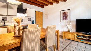 Un comedor con sillas y mesa de madera, un televisor sobre un mueble de madera, y una cocina al fondo.