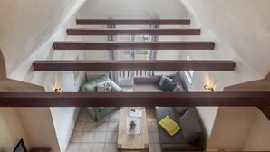 Un moderno espacio tipo loft con techo inclinado, vigas de madera y vista a una zona de estar acogedora abajo.