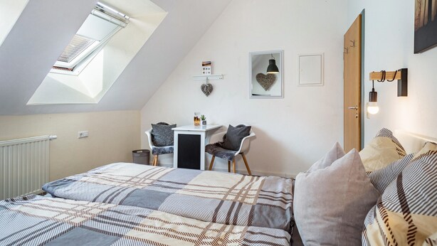 A bedroom with a skylight, a bed with patterned bedding, a small desk with chairs, and a wall-mounted shelf with decorative items. - Room 2, cozy, bright double room with sloping ceiling (Ravensburg)
